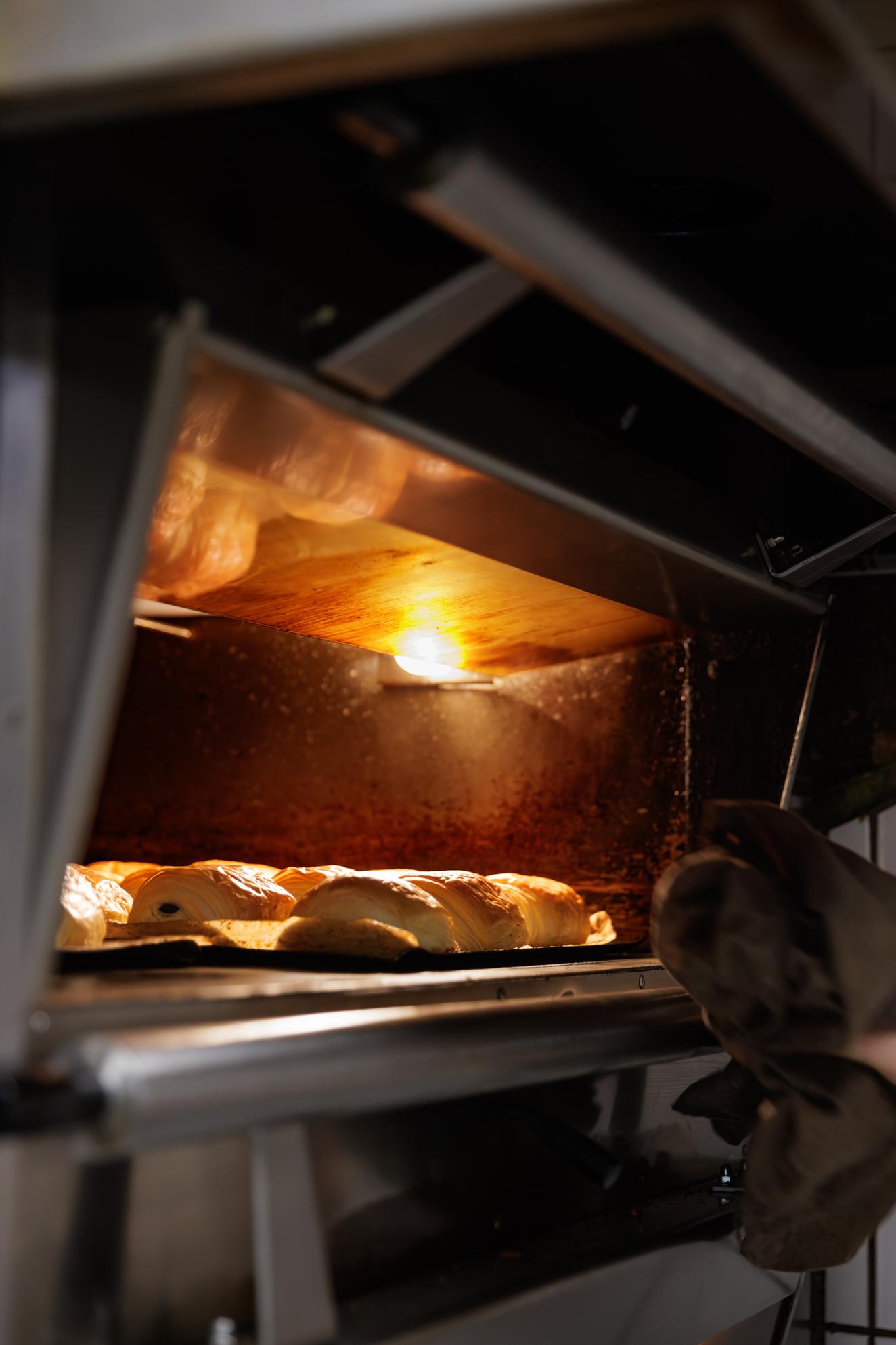 Main gantée enfournant une fournée de viennoiseries dans un four en lumière orangée.